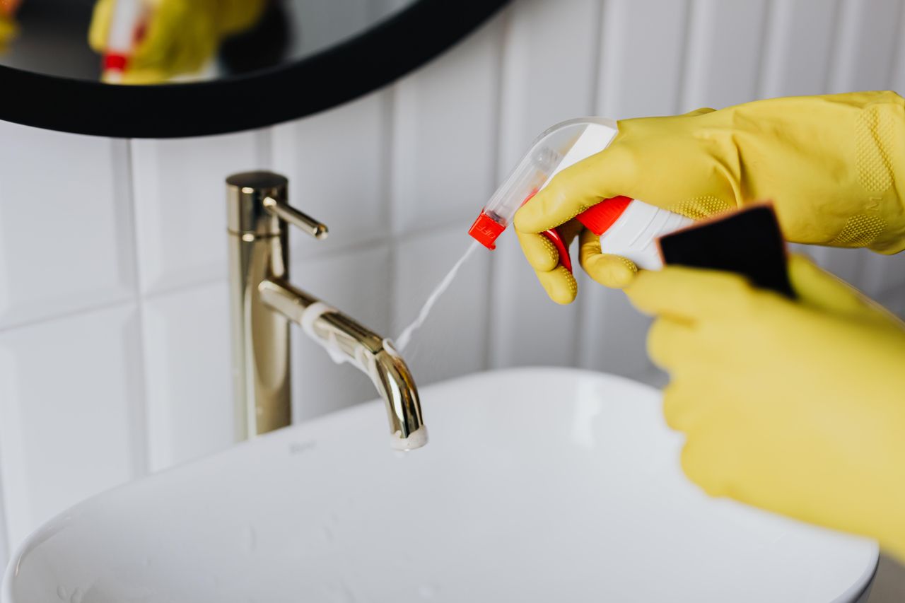 gallery-1 From above anonymous housekeeper in yellow rubber gloves spraying cleaner to chrome water faucet in bathroom